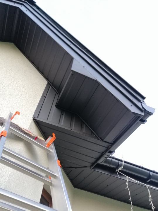 Fragment dachu z ciemnej blachy trapezowej, widoczna aluminiowa drabina oparta o ścianę budynku, rynna z elementami oświetlenia dekoracyjnego.