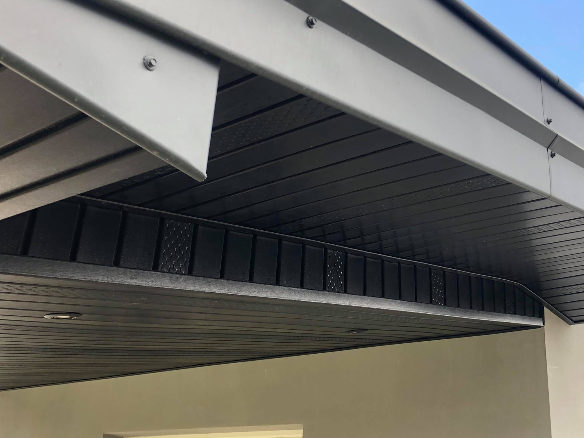 Czarny, nowoczesny okap dachowy z metalowymi obróbkami blacharskimi, widoczne wkręty mocujące i oświetlenie punktowe.