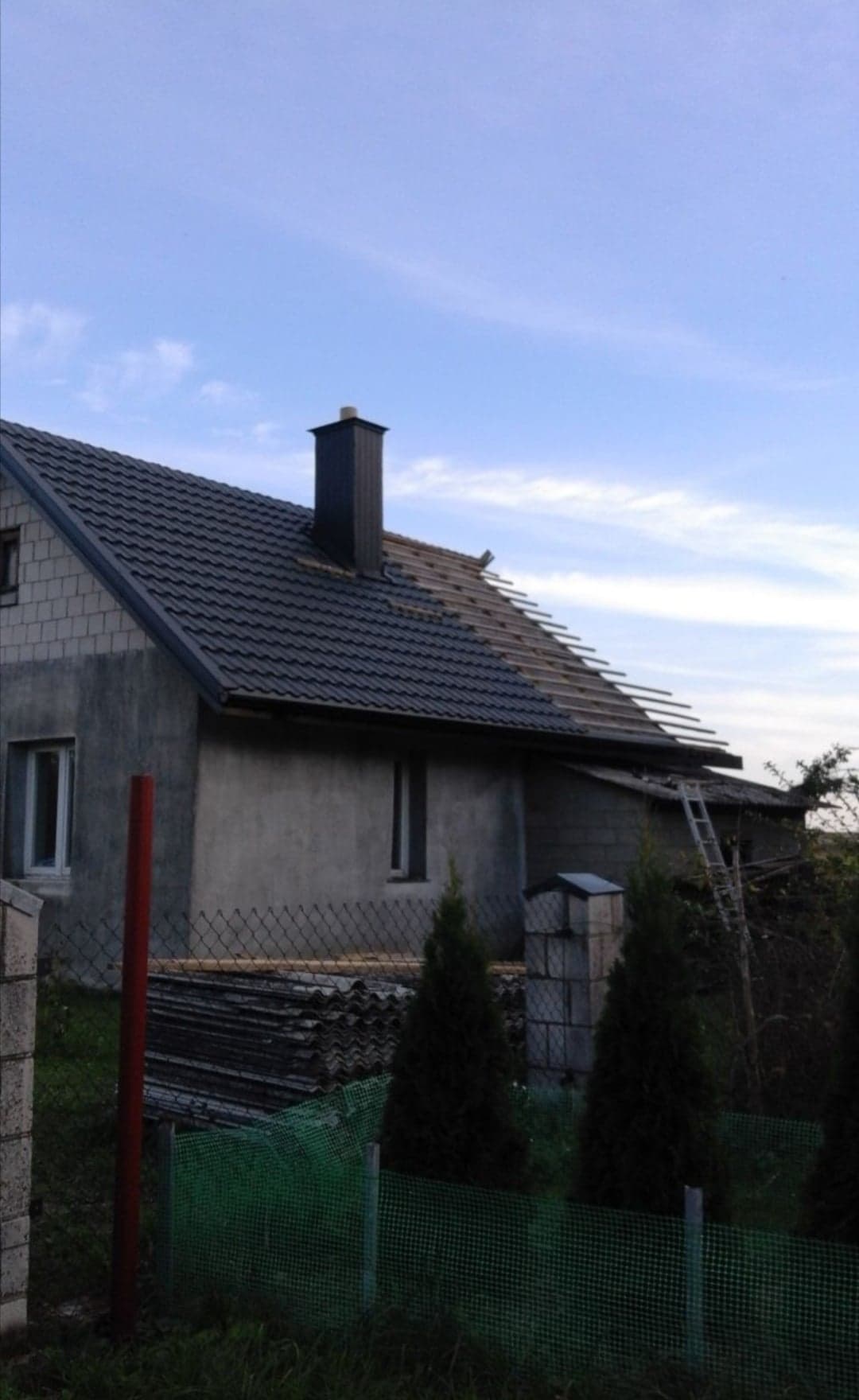 Dom jednorodzinny w trakcie wymiany pokrycia dachowego, widoczne nowe dachówki, komin z blachy i fragment starego dachu z eternitu, drabina oparta o budynek.