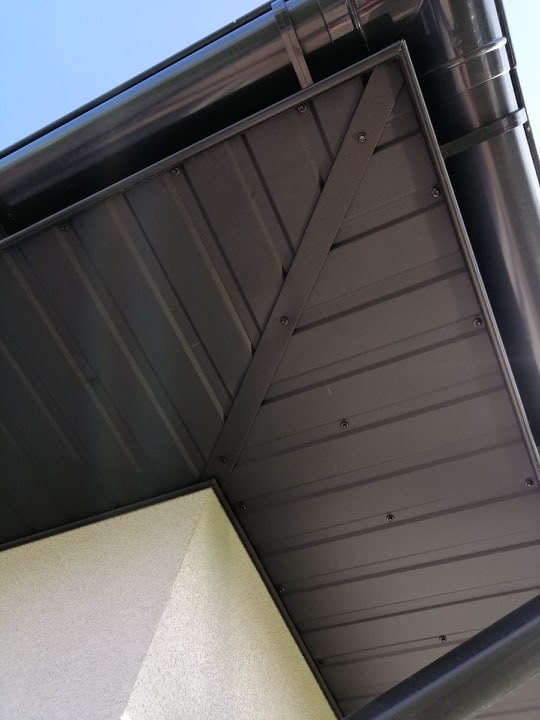 Szczegółowe ujęcie narożnika dachu z blachy trapezowej w kolorze grafitowym, widoczne mocowania i rynna, jasne tło ściany budynku.