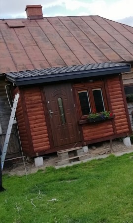 Drewniany ganek z nowym, ciemnym dachem blaszanym przylegający do starego domu z rdzawym dachem, drabina oparta o ścianę.