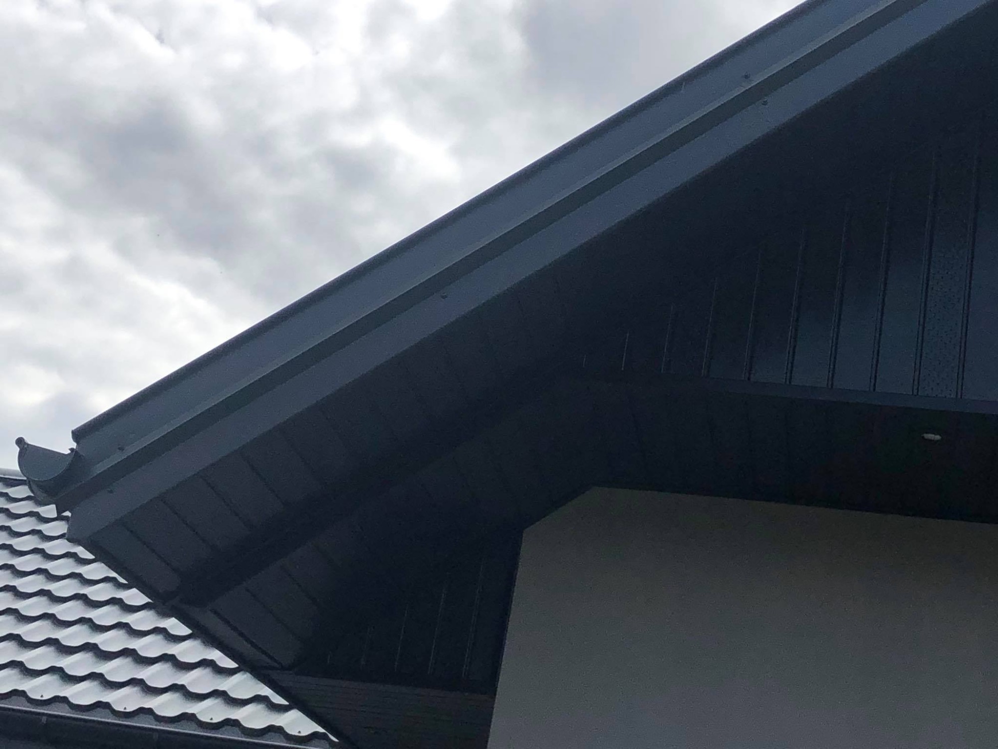 Fragment dachu z ciemną blachodachówką, obróbką blacharską i rynną pod pochmurnym niebem.