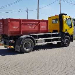 Żółta ciężarówka z czerwoną skrzynią transportową z napisem 'GRUZ ŚMIECI ODPADY' stoi na placu wyłożonym kostką brukową, w tle widoczna druga żółta ciężarówka Scania i ogrodzenie.