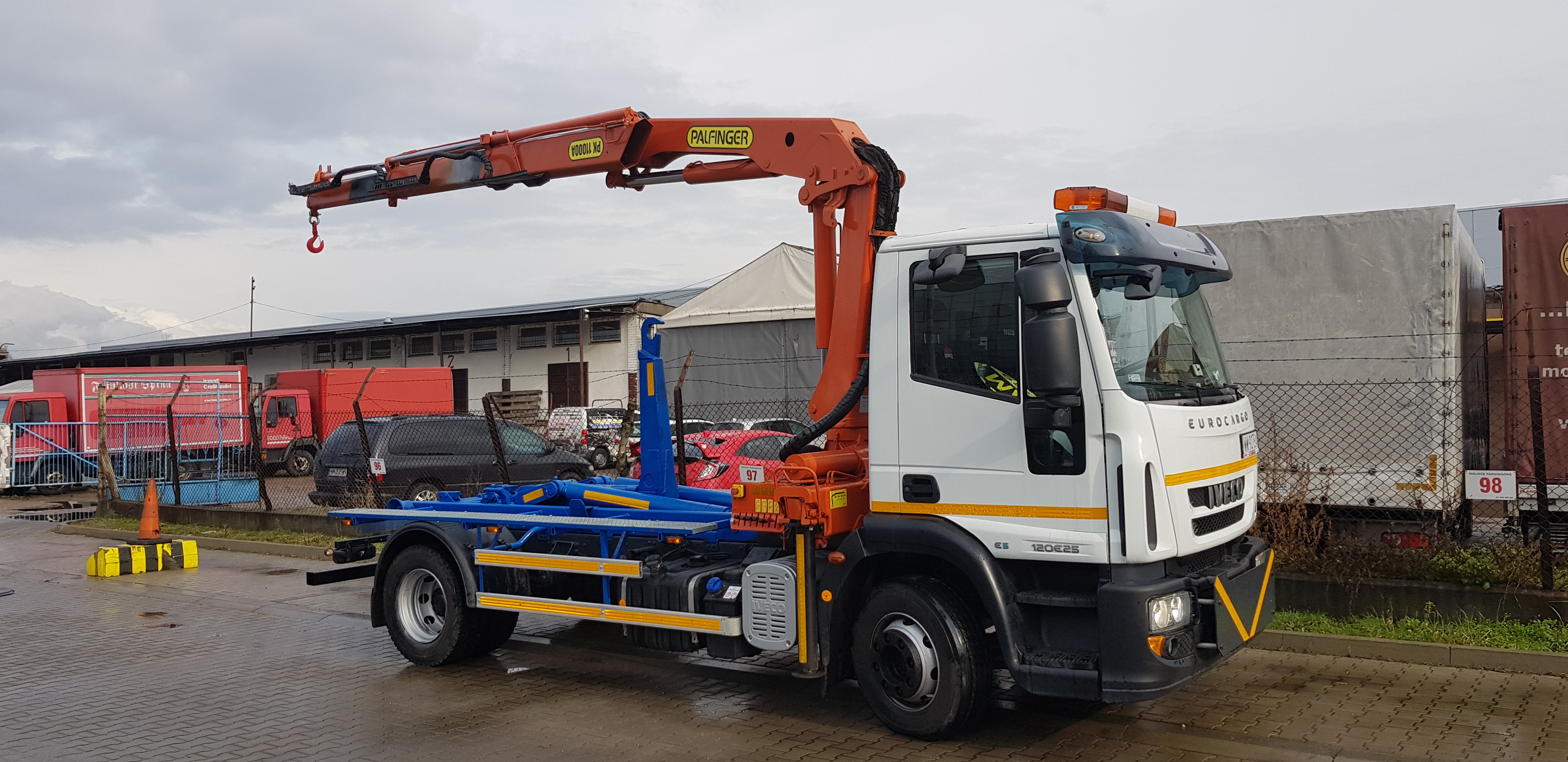 Ciężarówka Iveco Eurocargo z pomarańczowym dźwigiem Palfinger i niebieską konstrukcją do transportu kontenerów, zaparkowana na placu z widocznymi innymi pojazdami i budynkami w tle.