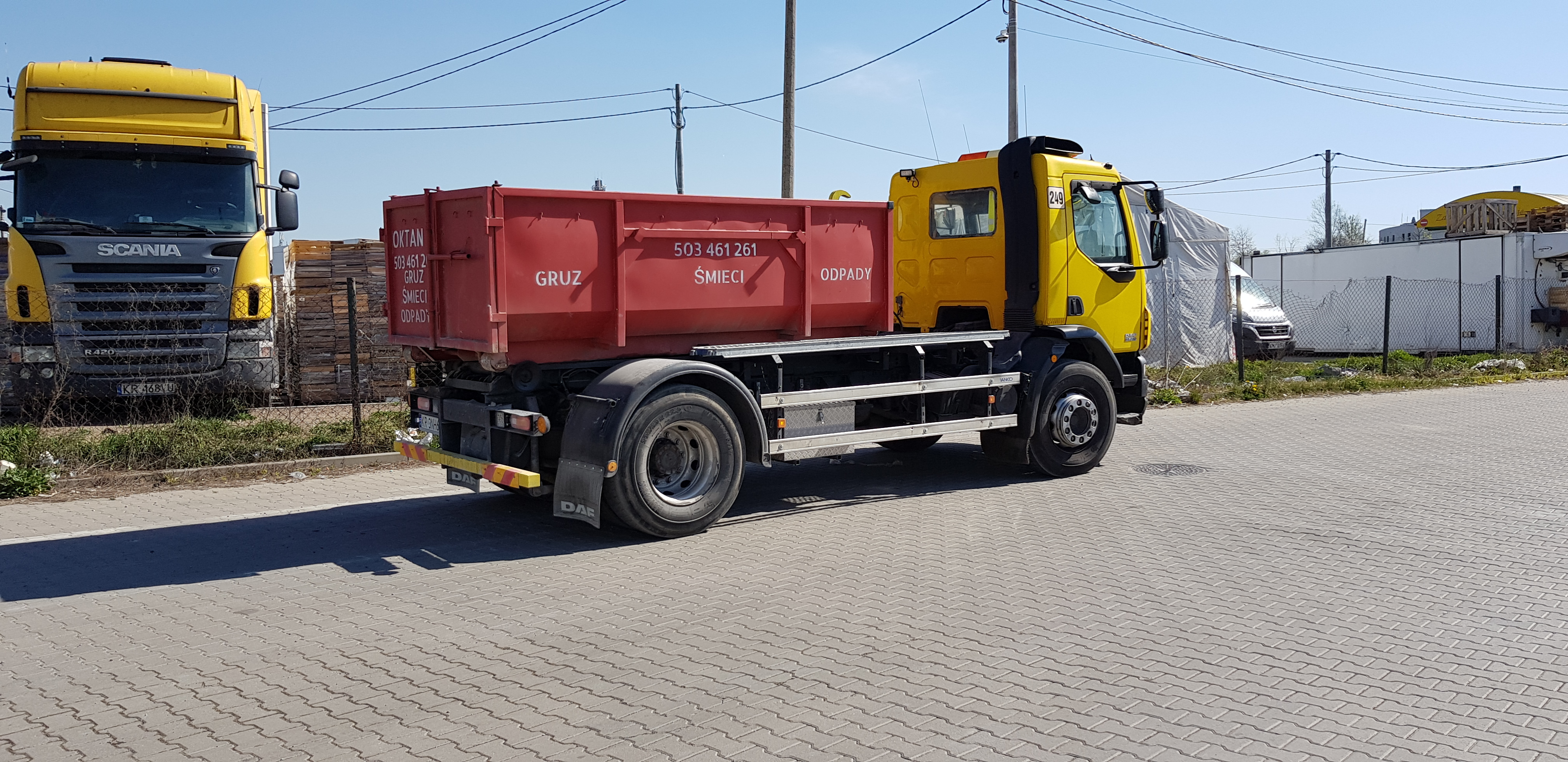 Żółta ciężarówka z czerwoną skrzynią transportową z napisem 'GRUZ ŚMIECI ODPADY' stoi na placu wyłożonym kostką brukową, w tle widoczna druga żółta ciężarówka Scania i ogrodzenie.
