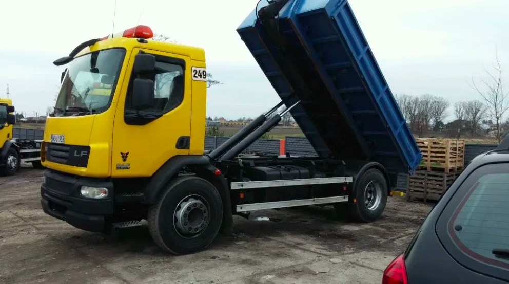 Żółta wywrotka DAF LF z podniesioną, niebieską skrzynią ładunkową na placu budowy, widoczne siłowniki hydrauliczne i palety w tle.