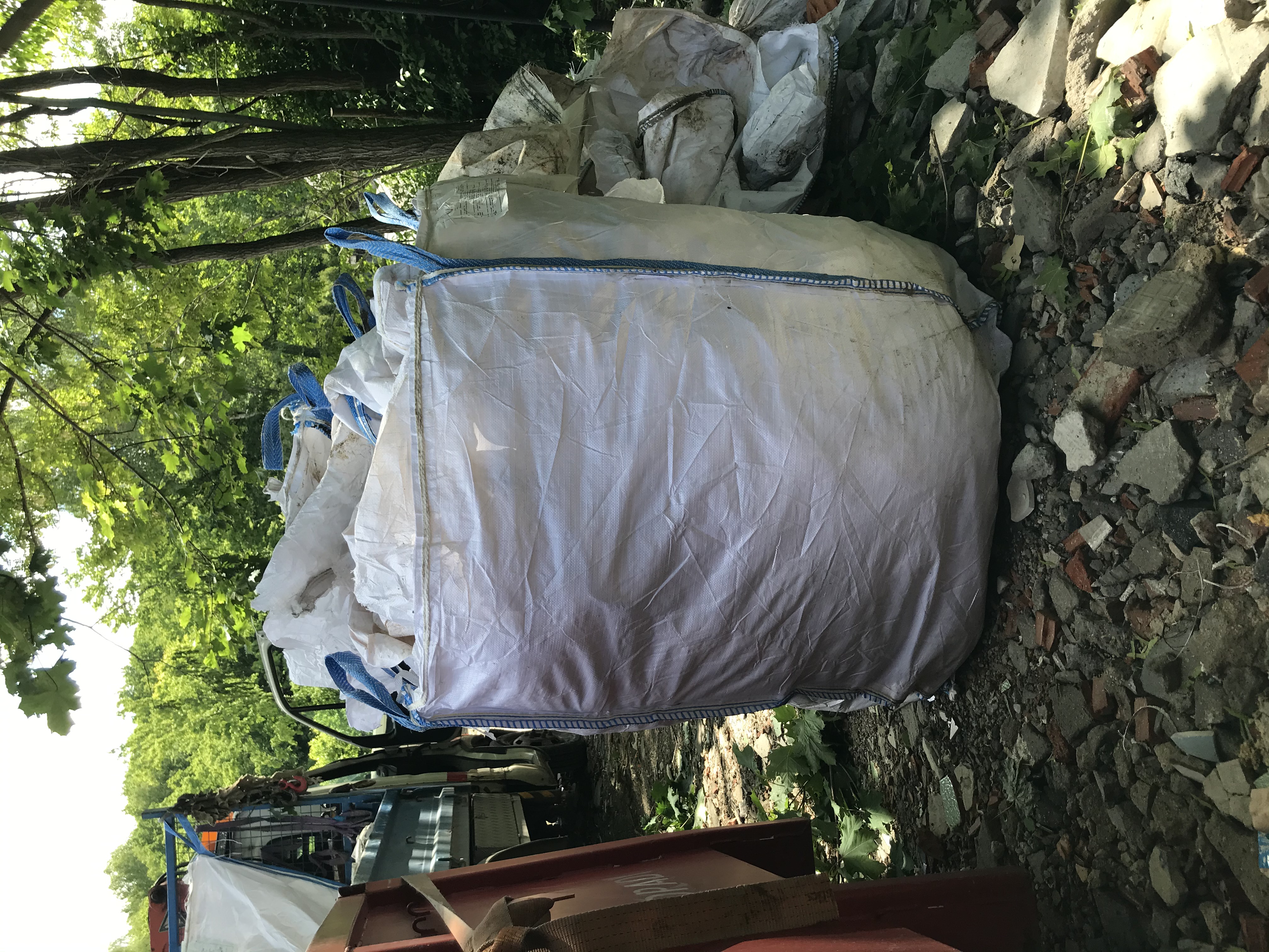 Duży, biały worek typu Big Bag z niebieskimi uchwytami, wypełniony gruzem i ustawiony na przyczepie samochodu ciężarowego, otoczony gruzem i zielenią.