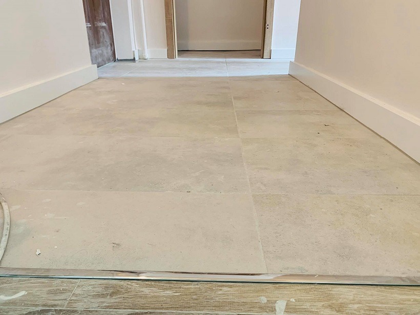 Korytarz z ułożoną jasną, kwadratową terakotą, białe listwy przypodłogowe, widoczne przejście do kolejnego pomieszczenia z surowym wykończeniem ościeżnicy.