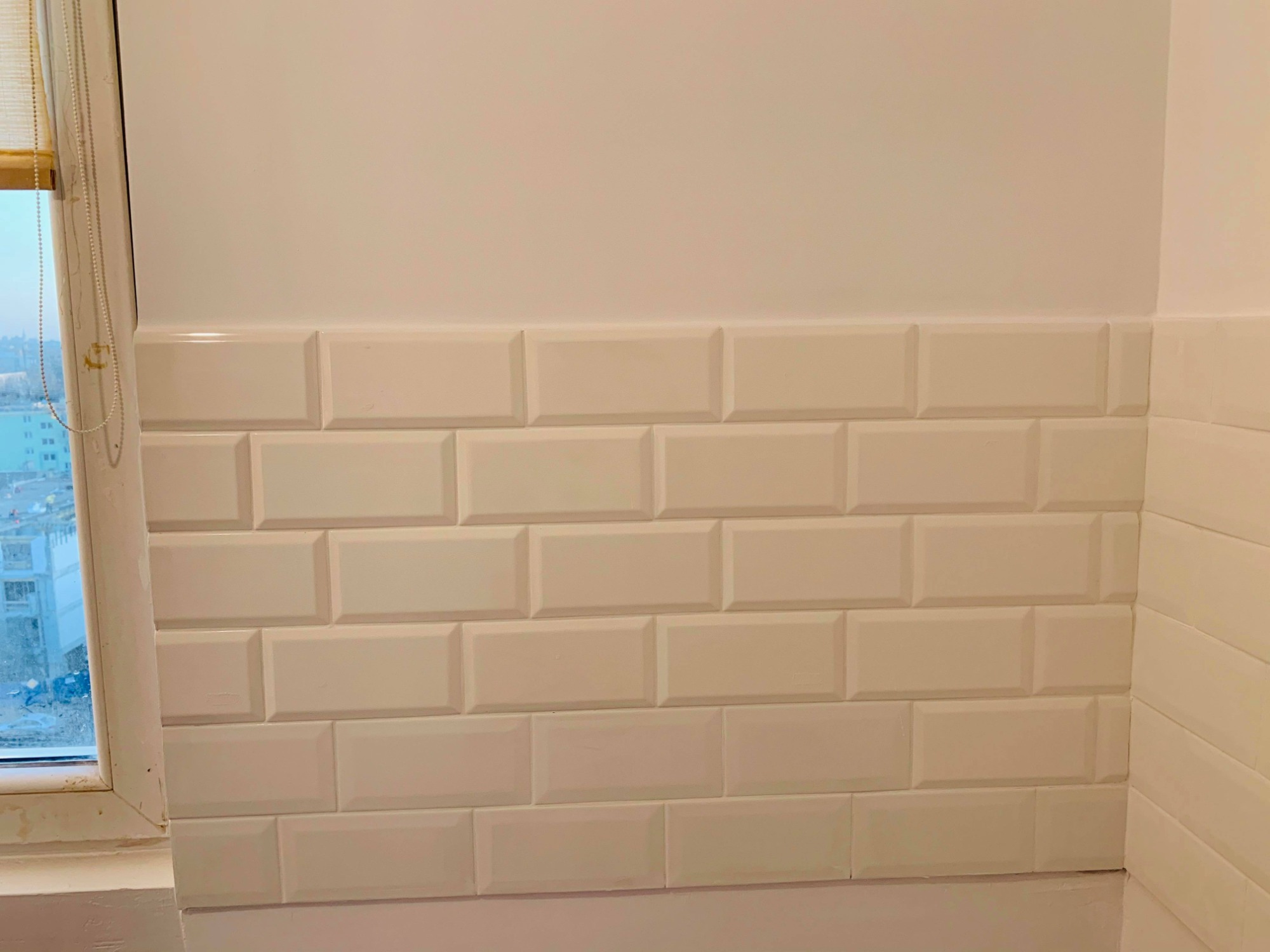 Ściana wyłożona białymi, wypukłymi płytkami ceramicznymi typu metro, z widocznym oknem po lewej stronie i fragmentem rolety.