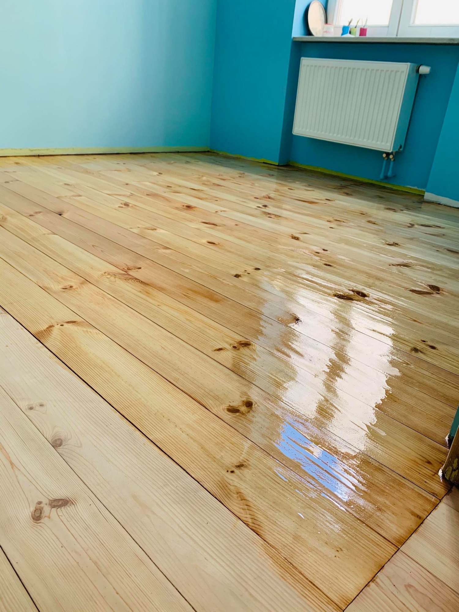 Świeżo polakierowana podłoga z jasnego drewna w pokoju z niebieskimi ścianami, widoczne refleksy światła i fragment grzejnika.