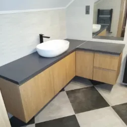 Łazienka na poddaszu z umywalką nablatową na blacie z szarego konglomeratu i szafkami z jasnego drewna, czarna bateria, podłoga w szachownicę, w lustrze odbija się grzejnik drabinkowy.