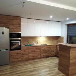 Nowoczesna kuchnia z drewnianymi frontami szafek dolnych i białymi górnymi, z drewnianą wyspą i widocznym fragmentem okna.