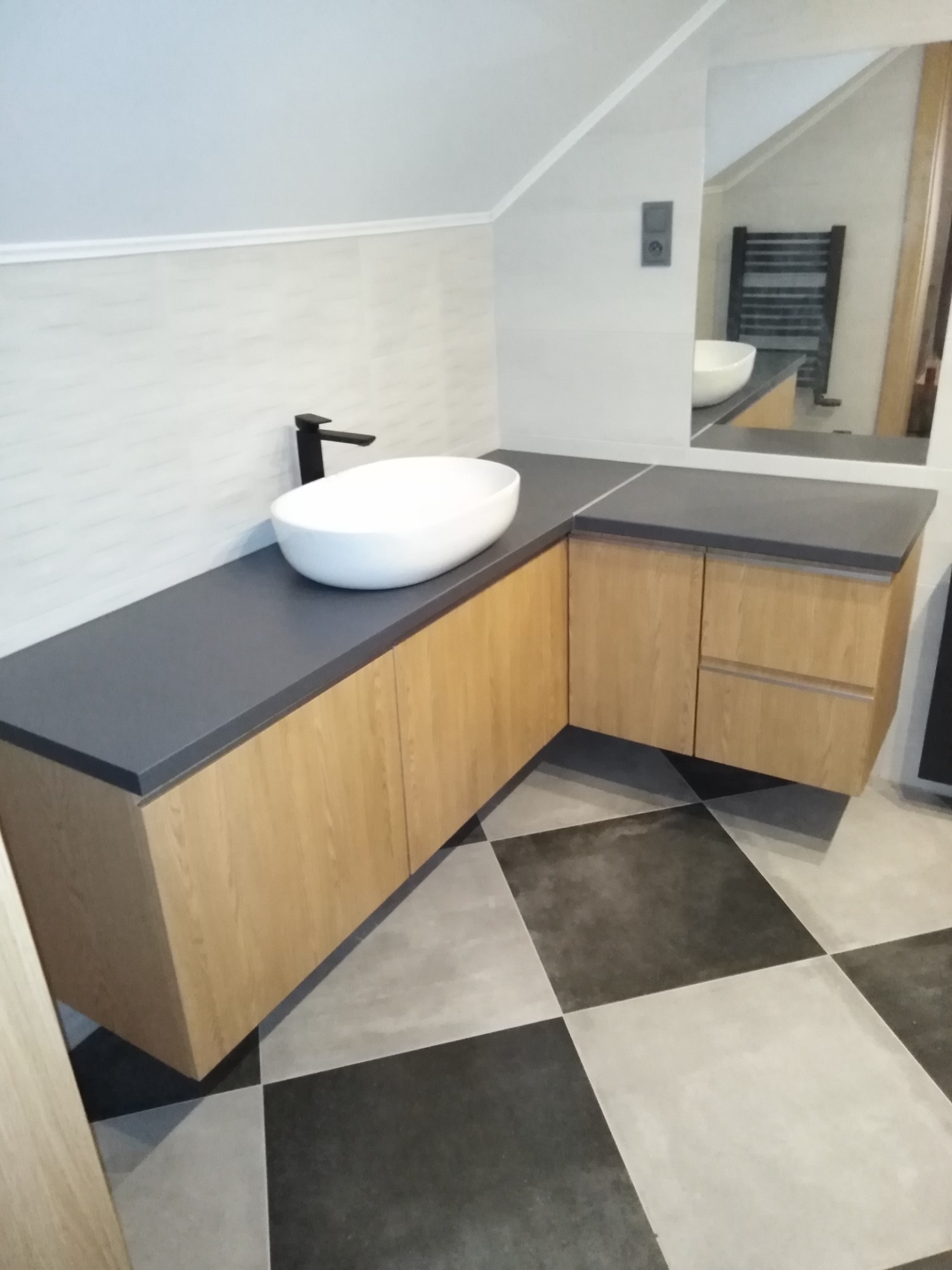 Łazienka na poddaszu z umywalką nablatową na blacie z szarego konglomeratu i szafkami z jasnego drewna, czarna bateria, podłoga w szachownicę, w lustrze odbija się grzejnik drabinkowy.