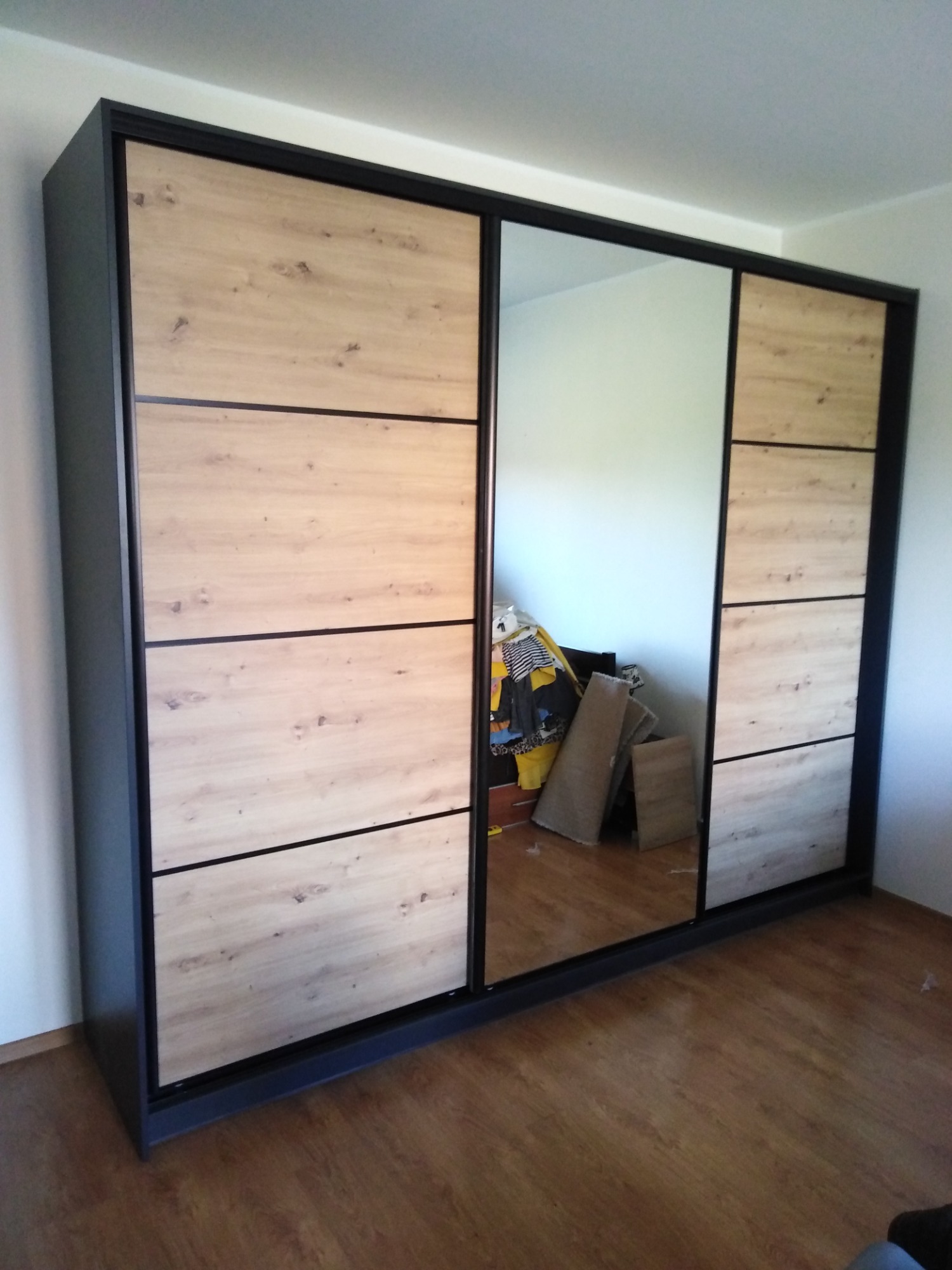 Nowoczesna szafa przesuwna z lustrem i drewnopodobnymi panelami, czarne obramowanie, widoczne odbicie wnętrza pokoju z ubraniami i elementami kartonowymi.
