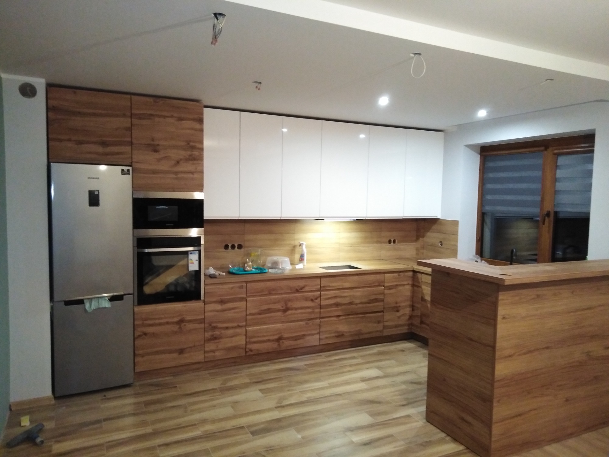 Nowoczesna kuchnia z drewnianymi frontami szafek dolnych i białymi górnymi, z drewnianą wyspą i widocznym fragmentem okna.