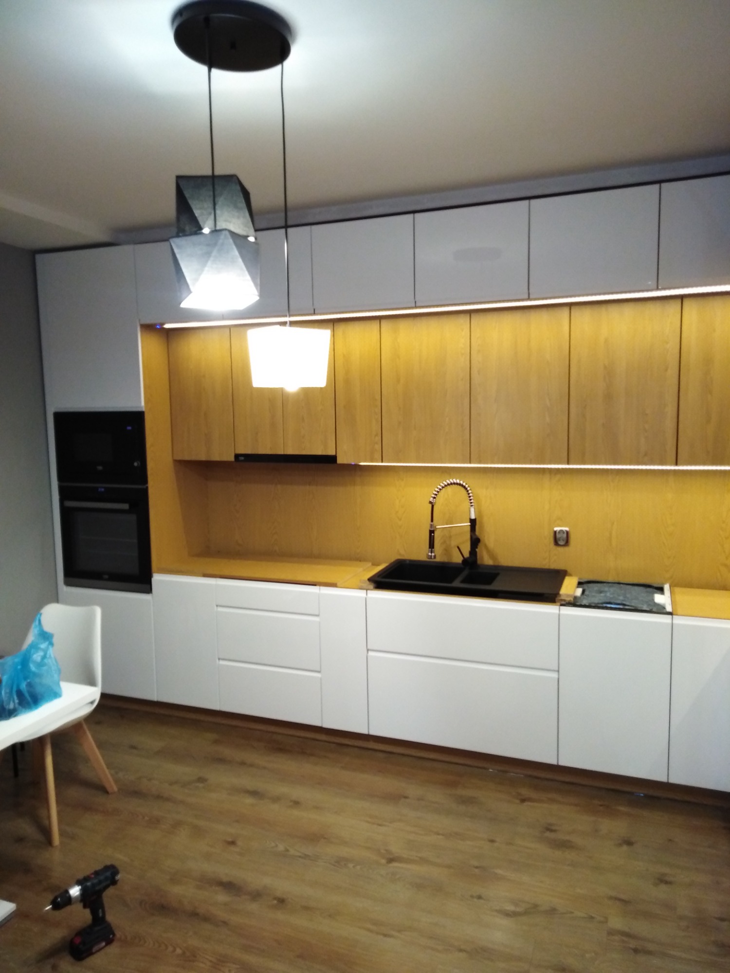Nowoczesna kuchnia z białymi szafkami dolnymi i drewnianymi górnymi, podświetlona listwą LED, z czarnym zlewozmywakiem i baterią, widoczna wiertarka akumulatorowa na podłodze.