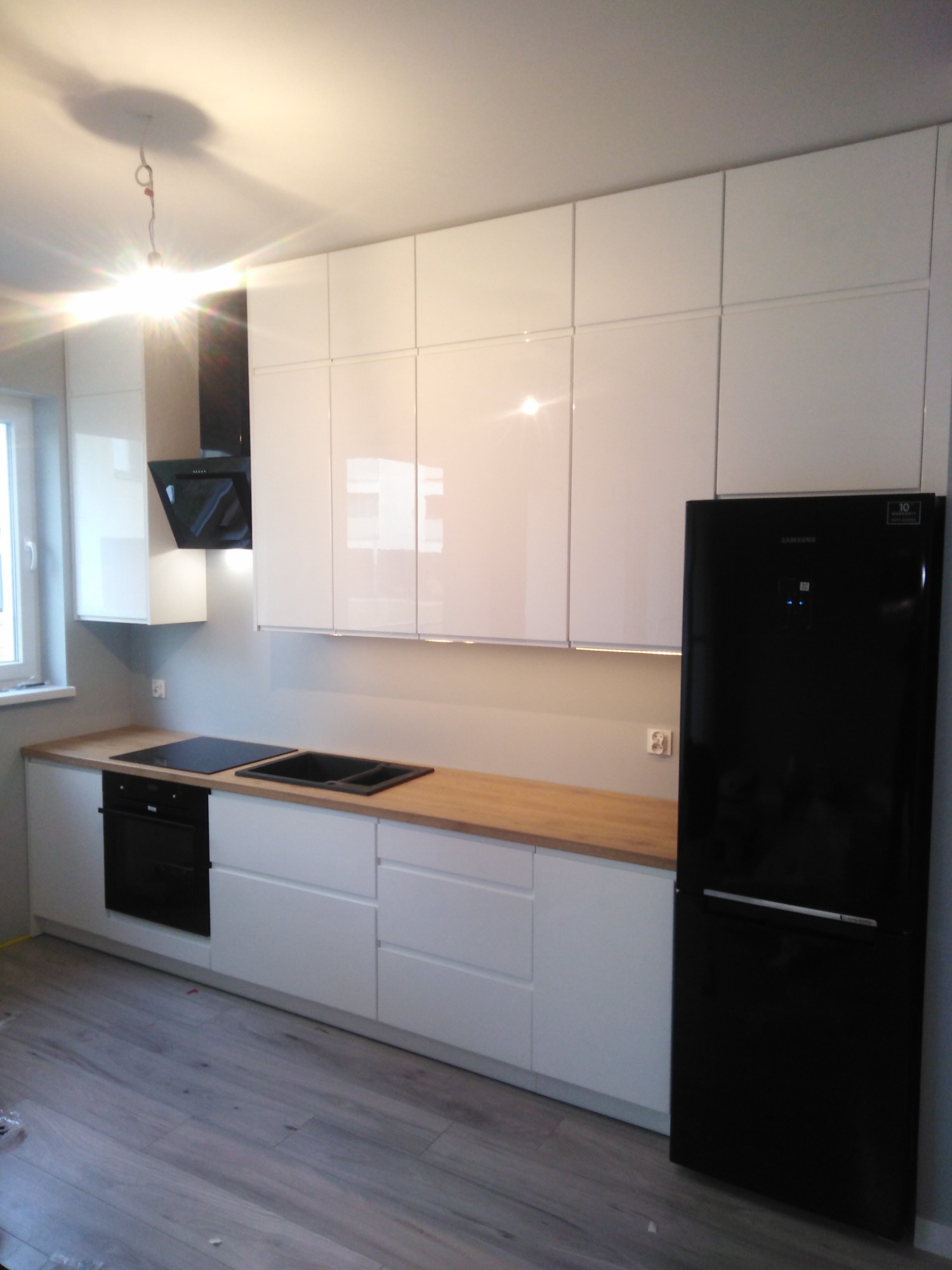 Nowoczesna kuchnia z białymi, lakierowanymi frontami, drewnianym blatem, czarną płytą indukcyjną i zlewem, oraz czarną lodówką Samsung, uchwycona z niskiego kąta.