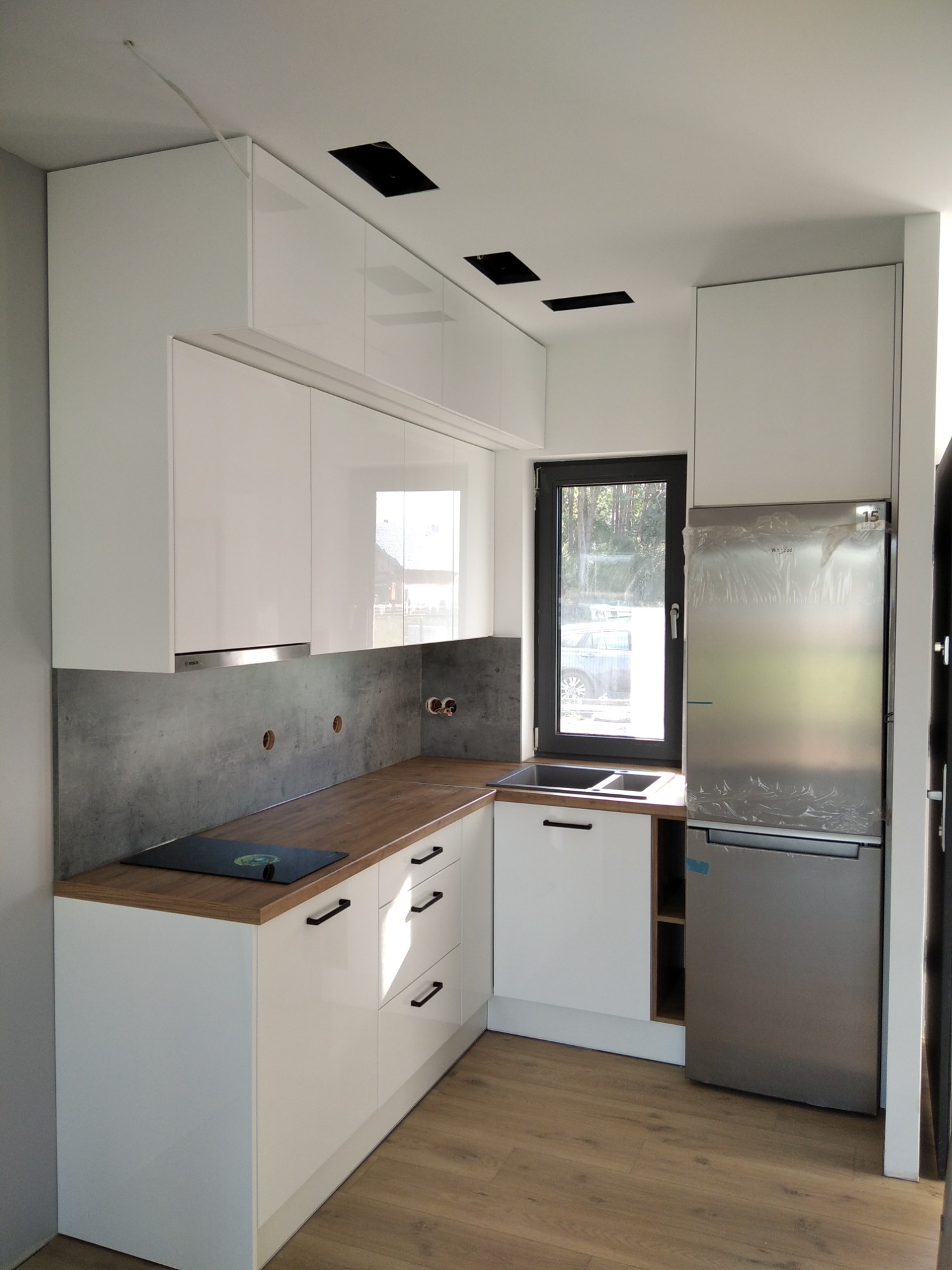 Nowoczesna, jasna kuchnia z białymi, lakierowanymi frontami szafek i drewnianym blatem. Uwagę zwraca szara ściana nad blatem oraz srebrna lodówka jeszcze w folii ochronnej. Widoczne są również...