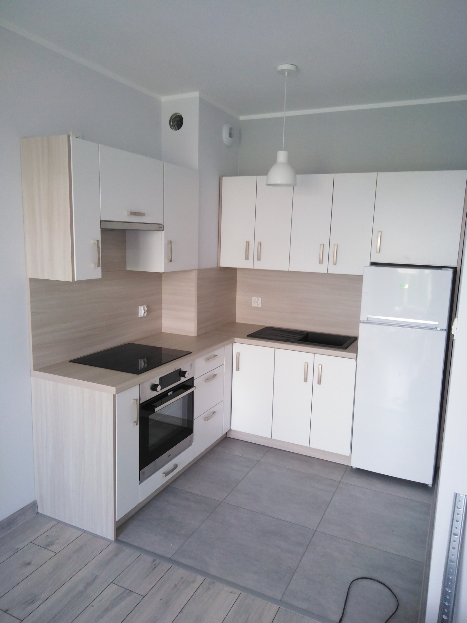 Nowoczesna, jasna kuchnia z białymi szafkami i drewnopodobnym blatem, płyta indukcyjna, piekarnik i biała lodówka, szare płytki podłogowe, minimalistyczna lampa wisząca.