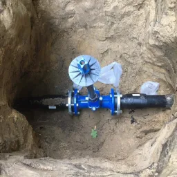 Złącze rury wodociągowej z niebieskim zaworem i kołnierzem w wykopie z piaszczystą ziemią, widoczne plastikowe worki.