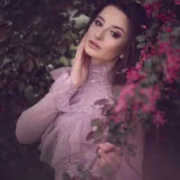 Portret kobiety w koronkowej sukni z falbanami, pozującej na tle kwitnącego drzewa z różowymi kwiatami, delikatny makijaż i dłoń dotykająca policzka.