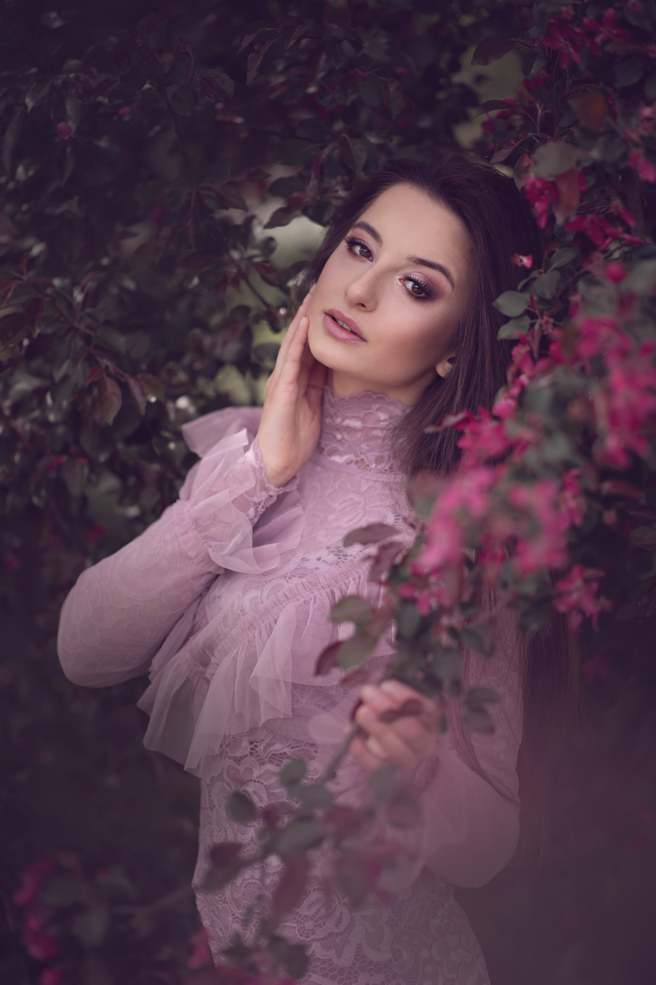 Portret kobiety w koronkowej sukni z falbanami, pozującej na tle kwitnącego drzewa z różowymi kwiatami, delikatny makijaż i dłoń dotykająca policzka.