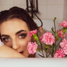Portret kobiety w wannie z mokrymi włosami, z makijażem oczu w odcieniach złota i brązu, otoczonej różowymi goździkami w paski.