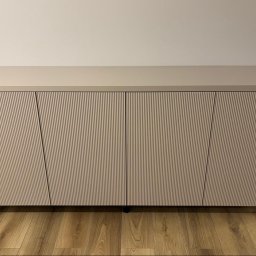 TW-MEBLE TOMASZ WOJTAŚ - Beżowa szafka RTV na kółkach z ryflowanymi frontami na drewnianej podłodze, widoczne gniazdko elektryczne i odkurzacz automatyczny.