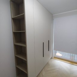 TW-MEBLE TOMASZ WOJTAŚ - Biała szafa z czarnymi, pionowymi uchwytami, z otwartą półką z boku, na podłodze parkiet ułożony w jodełkę francuską, widok z niskiego kąta.