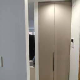 TW-MEBLE TOMASZ WOJTAŚ - Wysoka, dwudrzwiowa szafa wnękowa w odcieniach beżu z minimalistycznymi, podłużnymi uchwytami, widziana z perspektywy korytarza, z widocznym fragmentem wnętrza pokoju w tle.