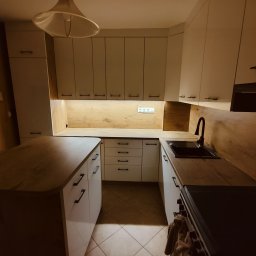 TW-MEBLE TOMASZ WOJTAŚ - Nowoczesna kuchnia z białymi szafkami i drewnianym blatem, czarna armatura i oświetlenie podszafkowe, widok na wyspę kuchenną i piekarnik z wiszącą szmatką.