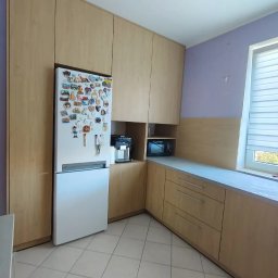 TW-MEBLE TOMASZ WOJTAŚ - Jasna kuchnia na wymiar z drewnianymi frontami, zabudowana lodówka z magnesami, szare blaty i płytki podłogowe.