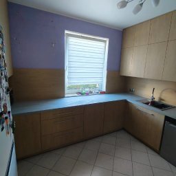 TW-MEBLE TOMASZ WOJTAŚ - Jasne drewniane szafki kuchenne z szarym blatem i białymi roletami w oknie, widok na fragment lodówki z magnesami i sufitową lampę.