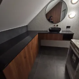 Łazienka na poddaszu z czarnym blatem i drewnianymi frontami szafek, owalnym lustrem i mozaiką na ścianie.