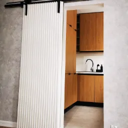 Białe, przesuwne drzwi z pionowymi żłobieniami na czarnym systemie szynowym, częściowo zasłaniające widok na drewniane szafki kuchenne i blat z czarnym zlewem.