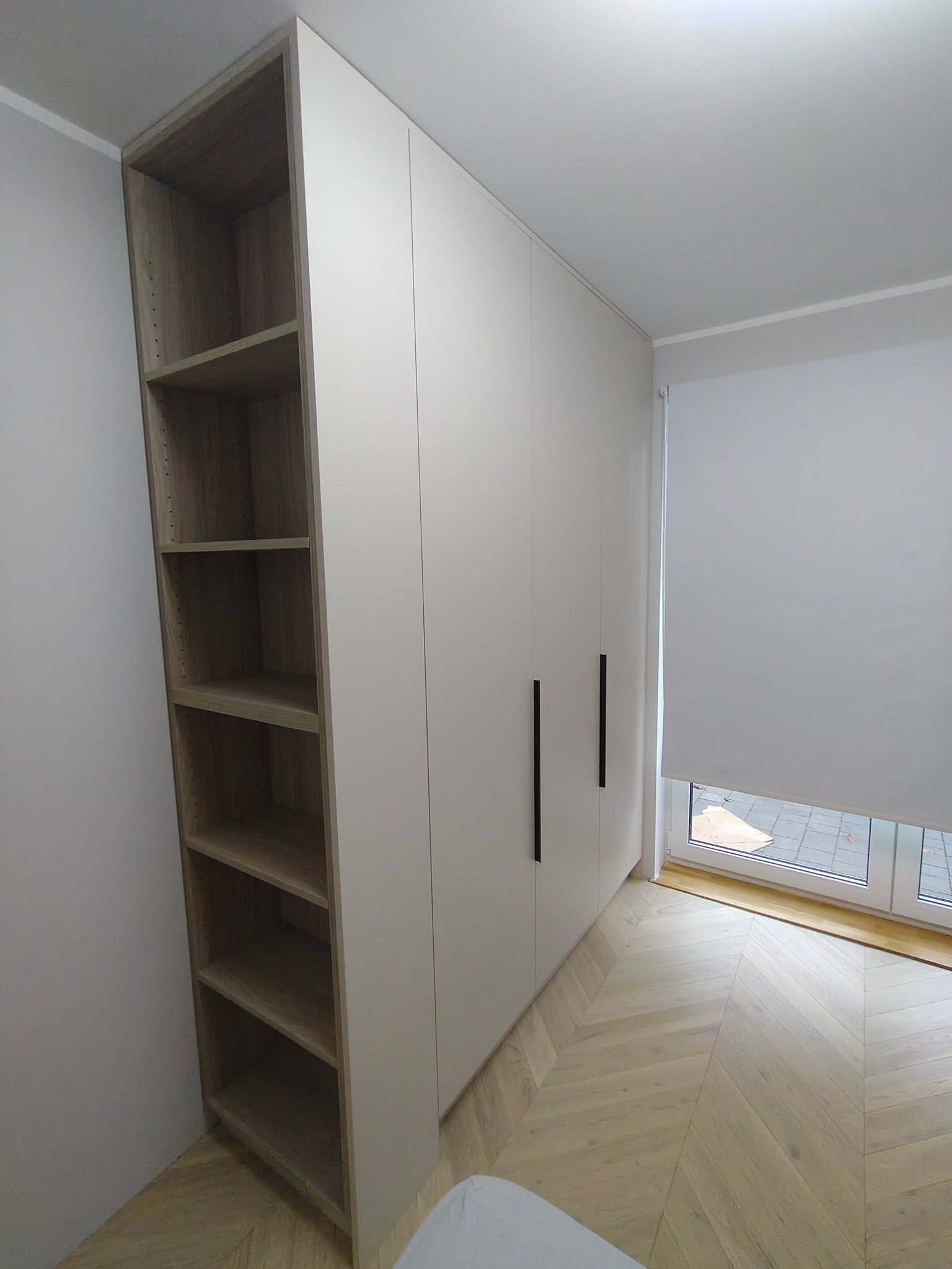 Biała zabudowa meblowa od podłogi do sufitu z otwartym regałem z drewnianymi półkami, widoczne czarne, pionowe uchwyty oraz podłoga ułożona w jodełkę.