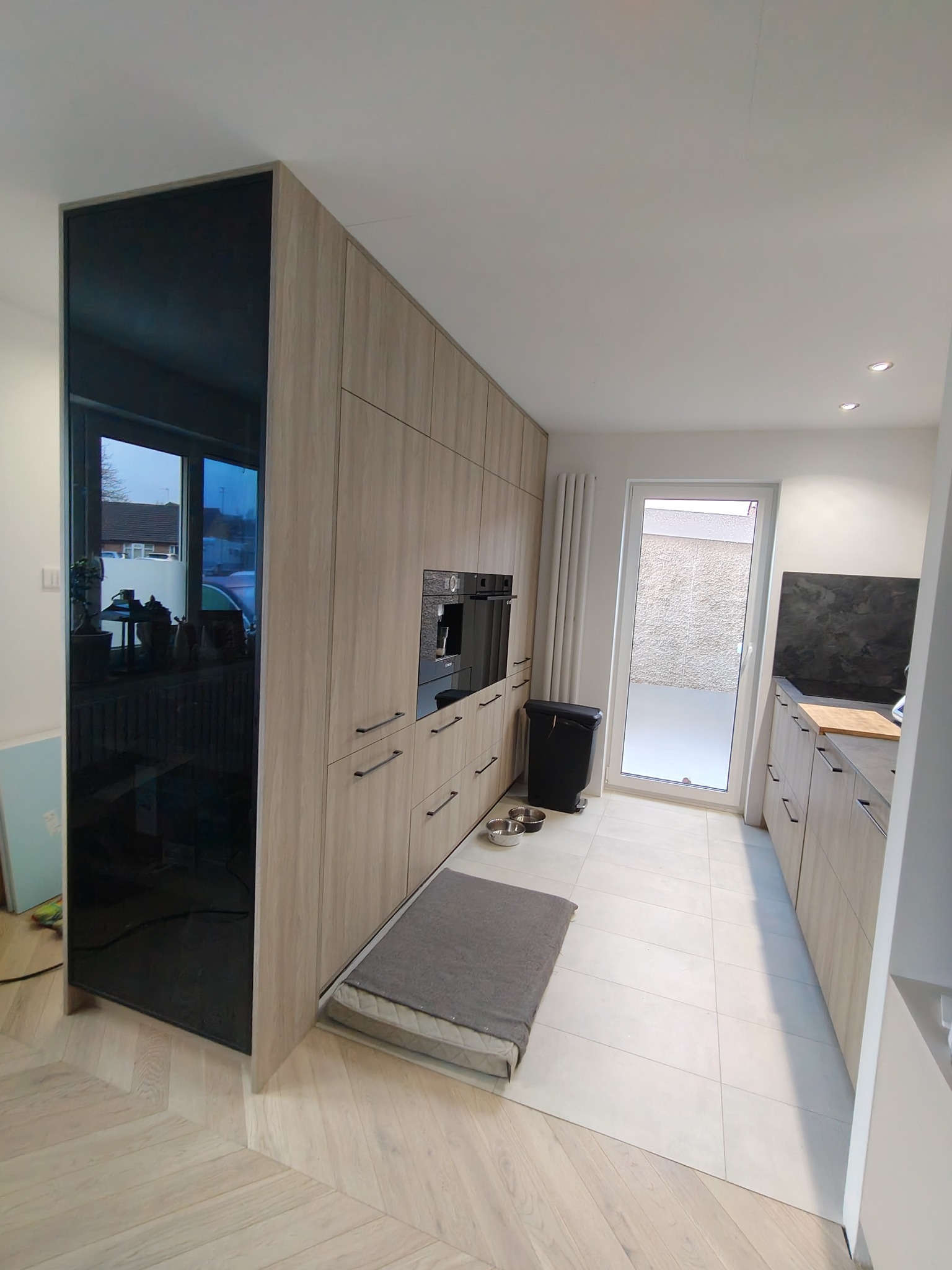 Nowoczesna kuchnia z zabudową meblową w odcieniach drewna, z czarnym panelem lustrzanym, zabudowanym ekspresem do kawy i widocznym legowiskiem dla psa