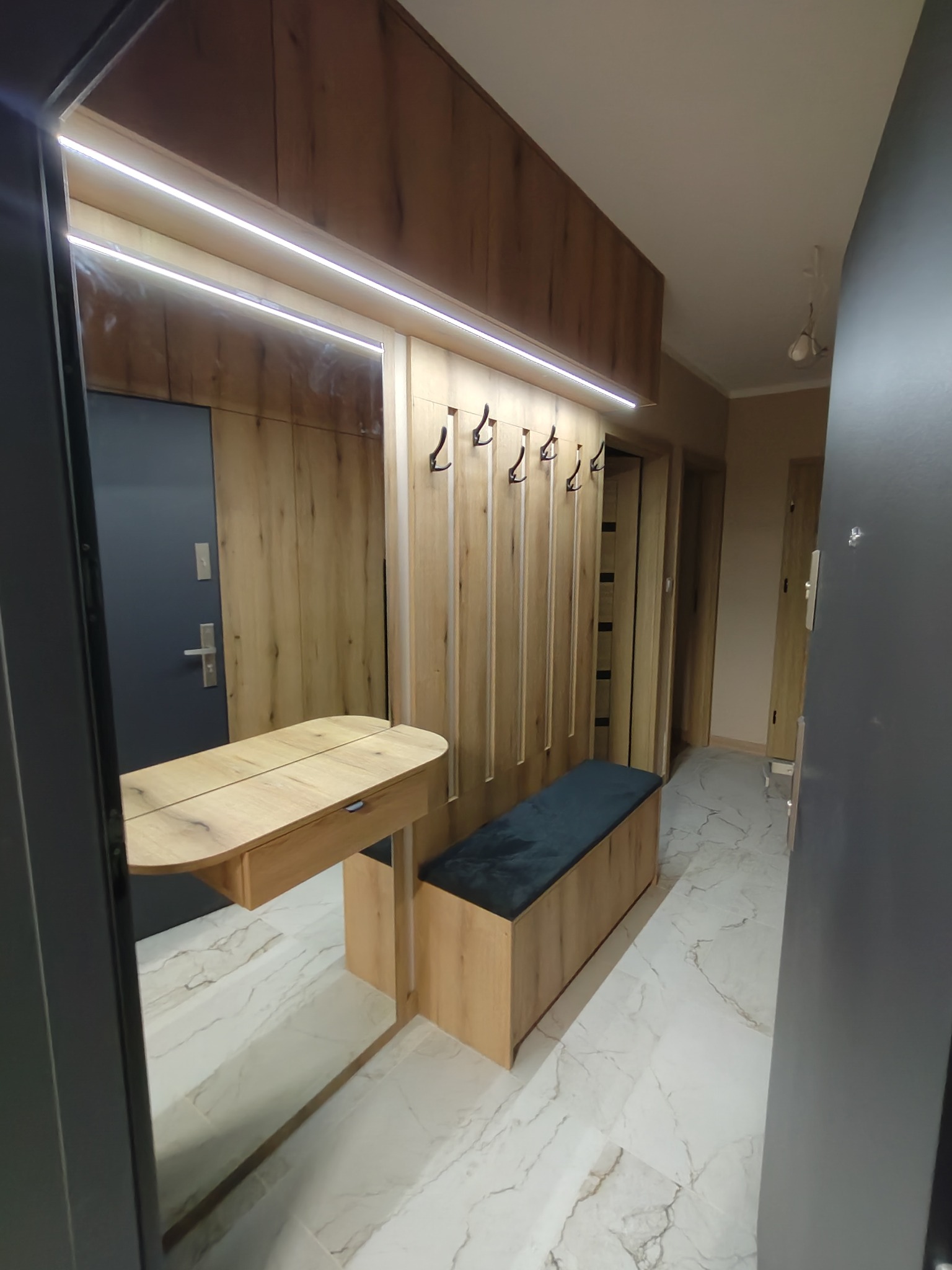 Wnętrze przedpokoju z drewnianą zabudową: szafki, wieszaki z haczykami, siedzisko z ciemnym obiciem i lustro z blatem. Całość oświetlona listwą LED. Podłoga wyłożona płytkami imitującymi marmur.