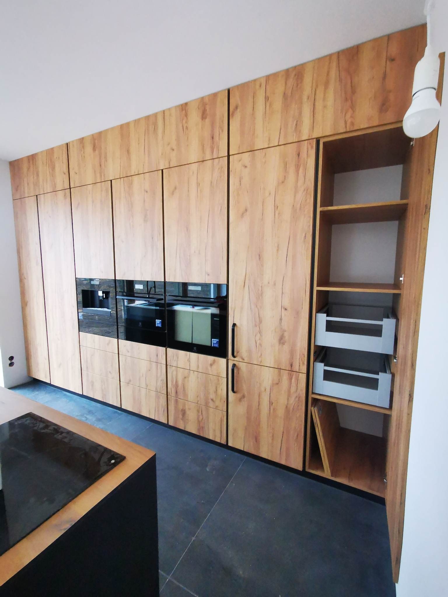 Nowoczesna zabudowa kuchenna z frontami w odcieniach drewna, z wbudowanymi urządzeniami i otwartą szafką z szufladami, oświetlona wiszącą żarówką.