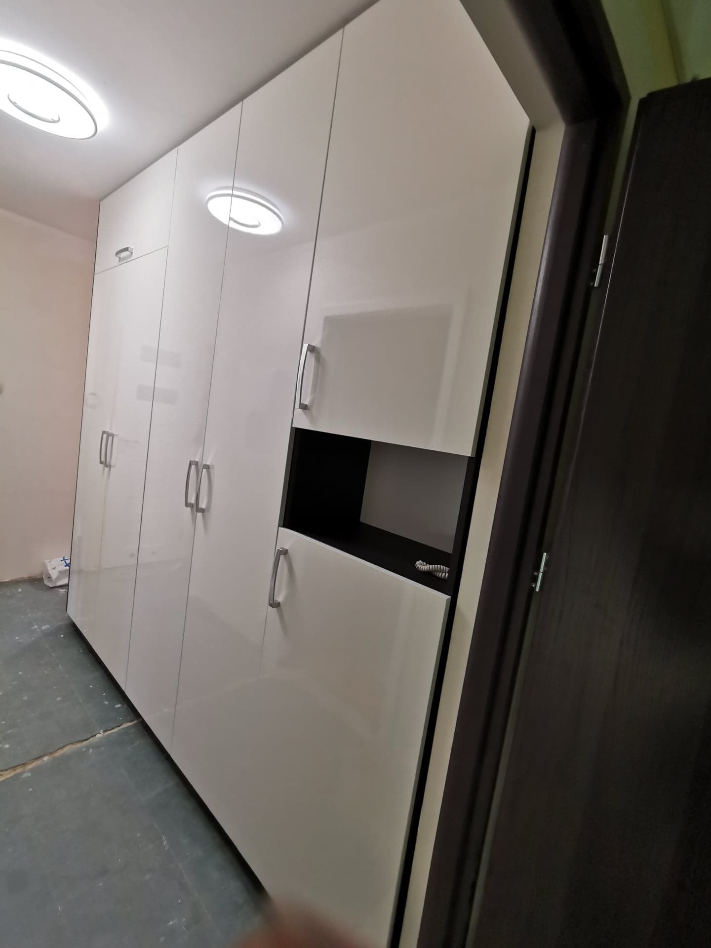 Biała, wysoka szafa z połyskiem, z otwartą półką z czarnym wnętrzem, widoczna przy wejściu do pokoju z ciemnymi drzwiami.