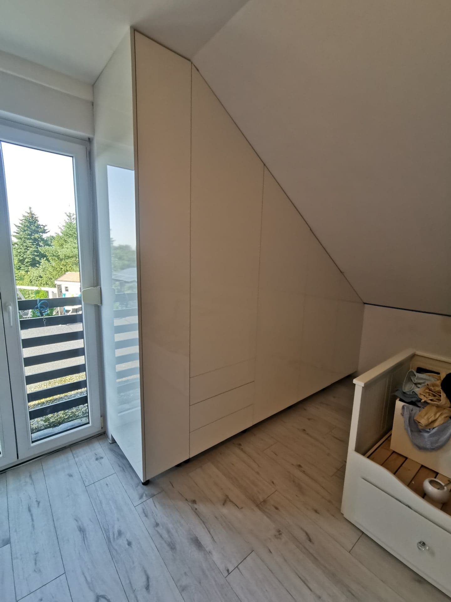 Biała szafa wnękowa na poddaszu w Poznaniu, dopasowana do skosu dachu, z widocznym oknem balkonowym i jasną podłogą imitującą drewno.