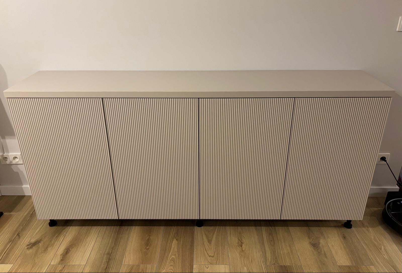 Beżowa szafka RTV na kółkach z ryflowanymi frontami na drewnianej podłodze, widoczne gniazdko elektryczne i odkurzacz automatyczny.