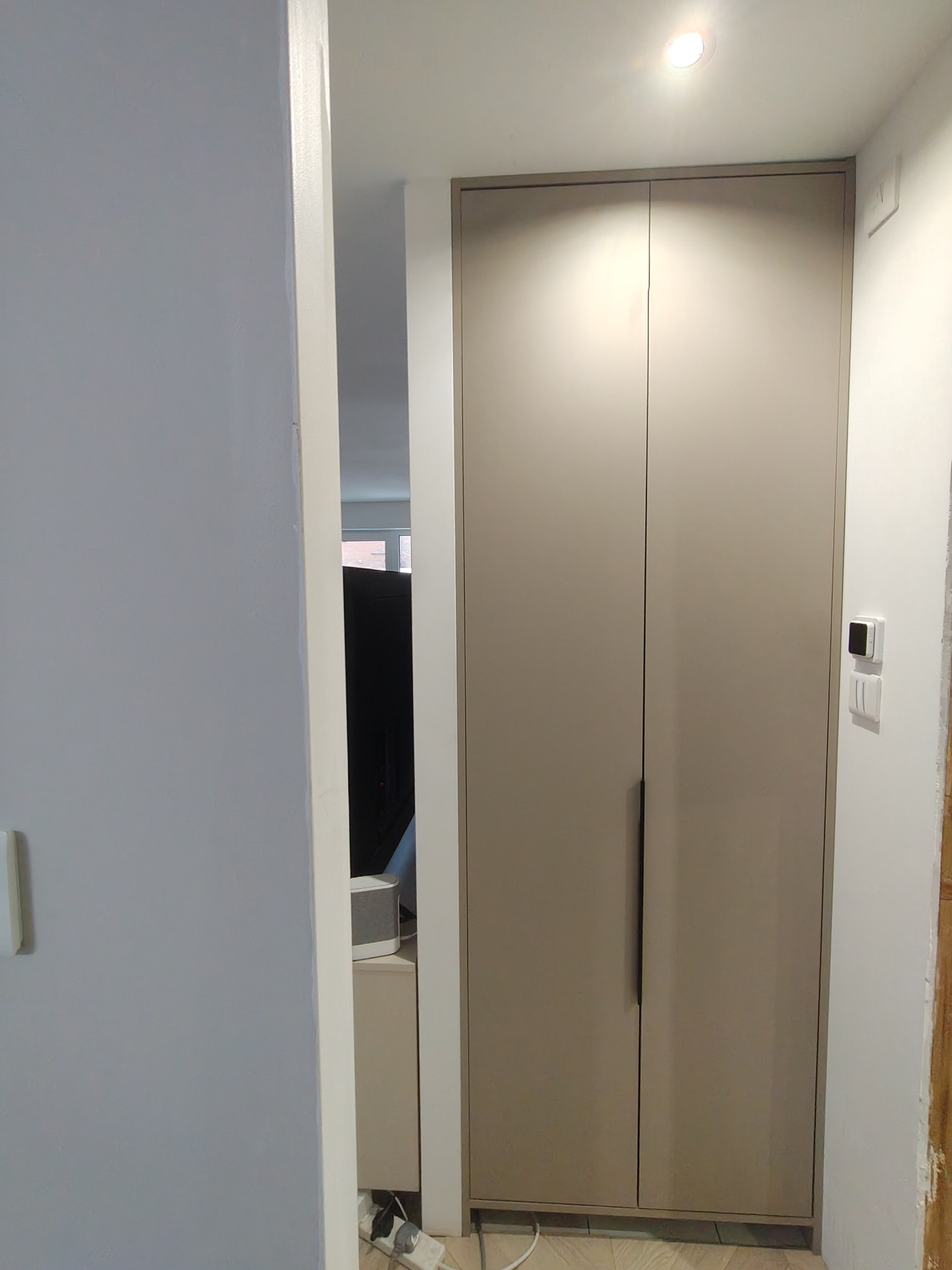 Wysoka, dwudrzwiowa szafa wnękowa w odcieniach beżu z minimalistycznymi, podłużnymi uchwytami, widziana z perspektywy korytarza, z widocznym fragmentem wnętrza pokoju w tle.