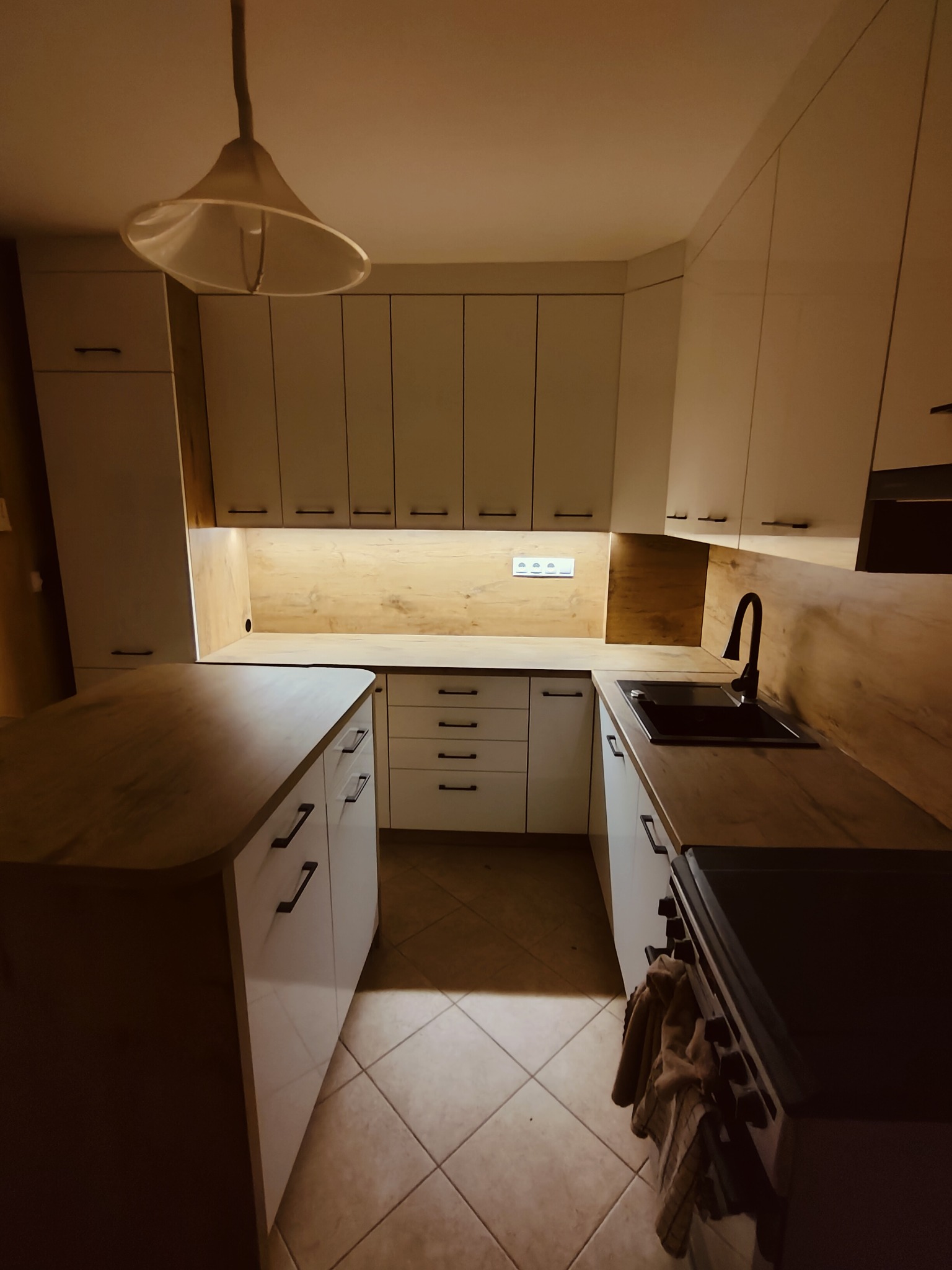 Nowoczesna kuchnia z białymi szafkami i drewnianym blatem, czarna armatura i oświetlenie podszafkowe, widok na wyspę kuchenną i piekarnik z wiszącą szmatką.