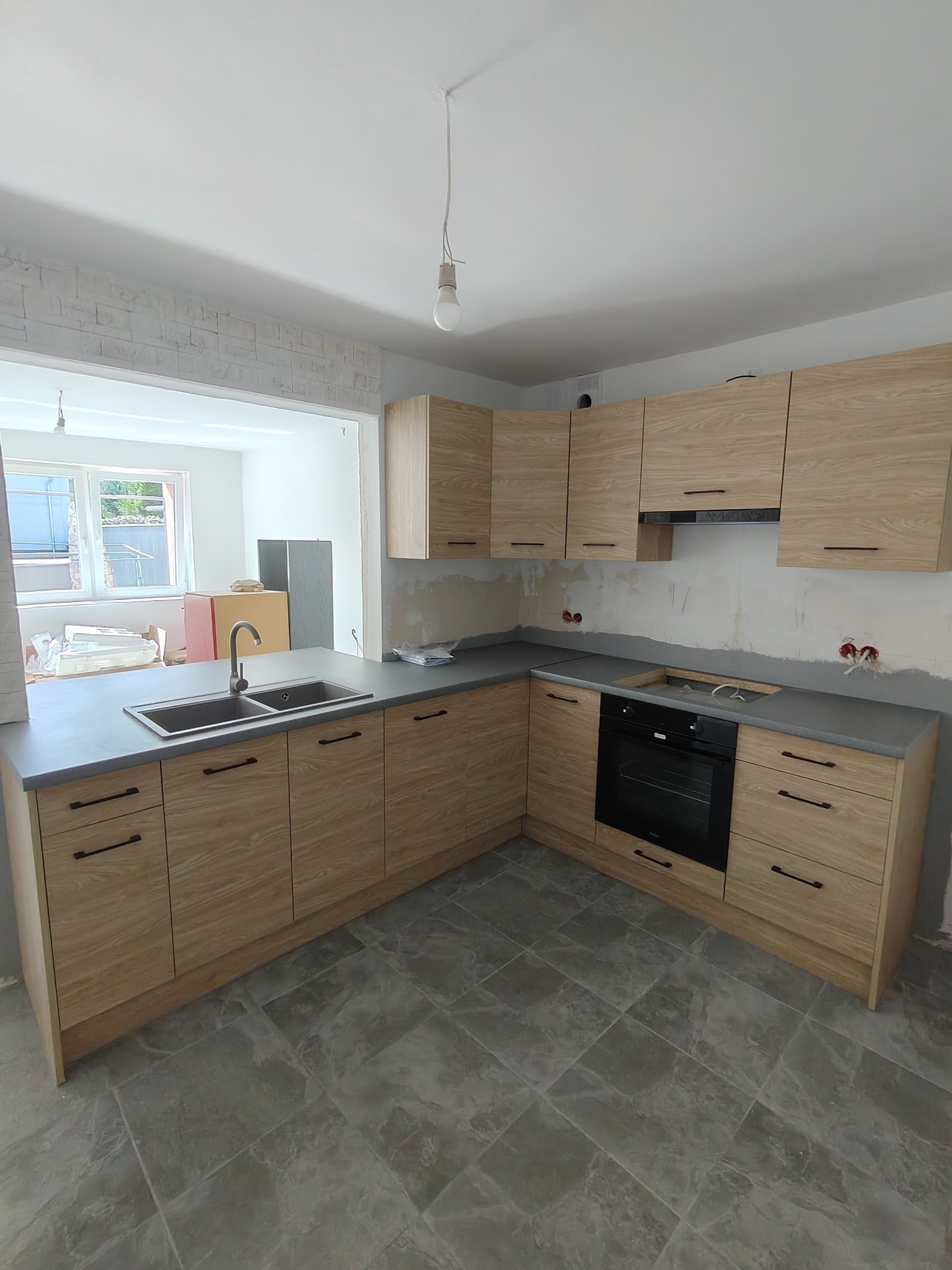 Kątowe ujęcie montażu jasnej zabudowy kuchennej z drewnopodobnymi frontami i szarym blatem, widoczne niezabezpieczone kable, surowa ściana i szare płytki podłogowe.