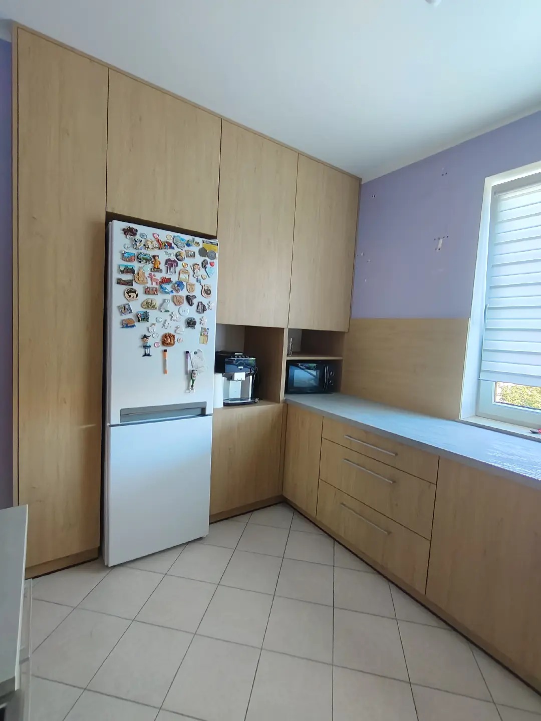 Jasna kuchnia na wymiar z drewnianymi frontami, zabudowana lodówka z magnesami, szare blaty i płytki podłogowe.