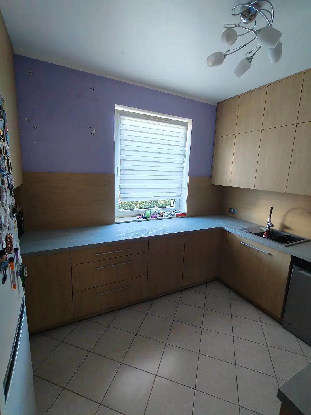 Jasne drewniane szafki kuchenne z szarym blatem i białymi roletami w oknie, widok na fragment lodówki z magnesami i sufitową lampę.