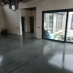 Gładka, szara posadzka w surowym wnętrzu z widoczną instalacją elektryczną na ścianie i dużymi oknami przesuwnymi, przez które widać teren budowy.