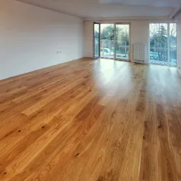 Jasne, drewniane deski podłogowe ułożone w przestronnym, pustym pokoju z białymi ścianami i dużymi oknami balkonowymi z widokiem na drzewa.