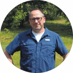 Portret mężczyzny w okularach i niebieskiej koszuli, z identyfikatorem na piersi, na tle zieleni.